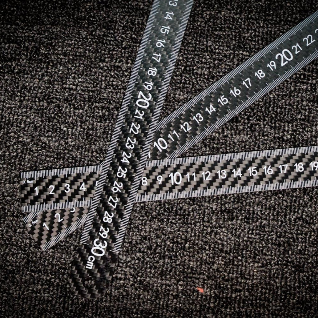 Three carbon fiber rulers with clear measurement markings, placed on a textured dark surface.