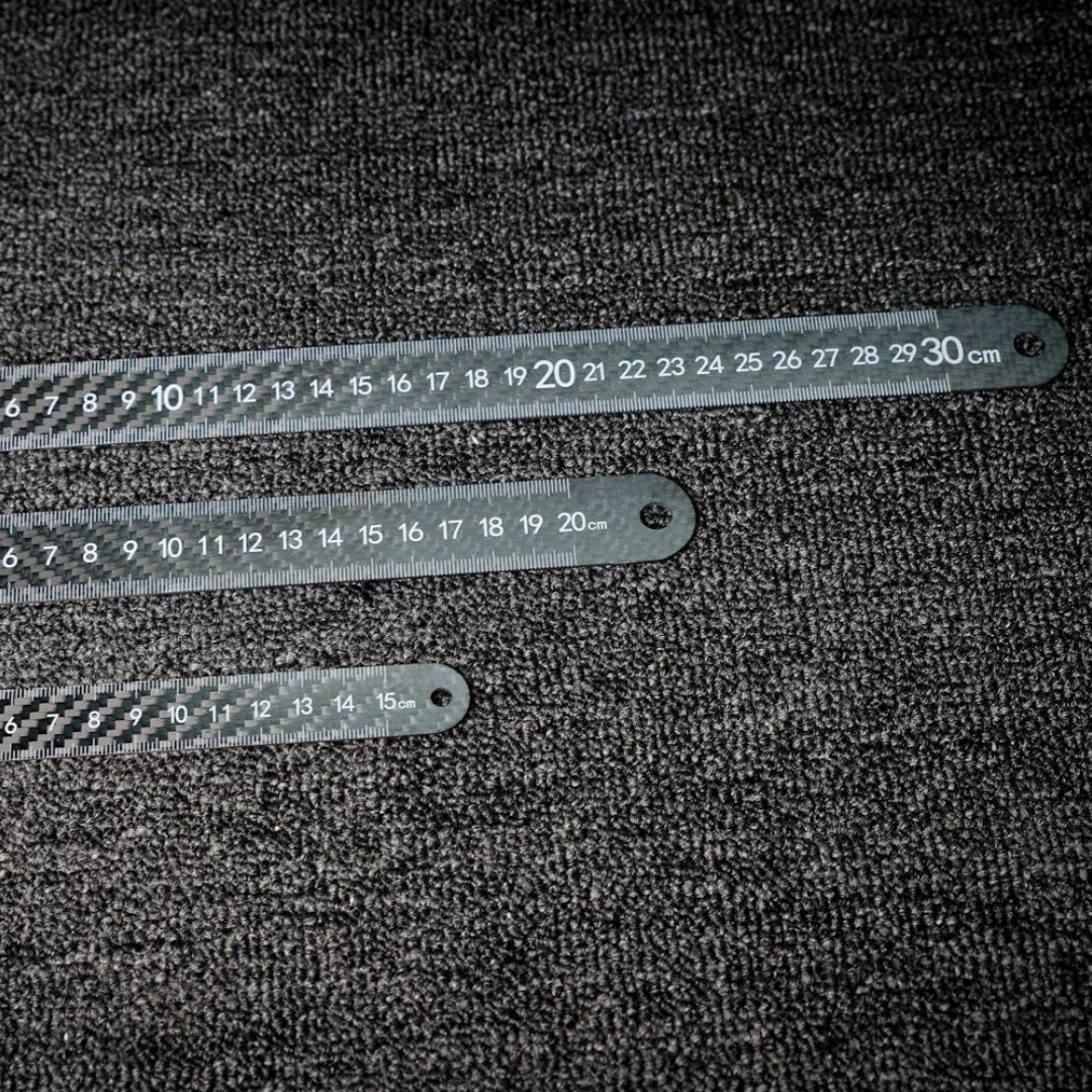 Three 3K carbon fiber rulers with distinct lengths and clear measurement markings, placed on a dark textured surface.