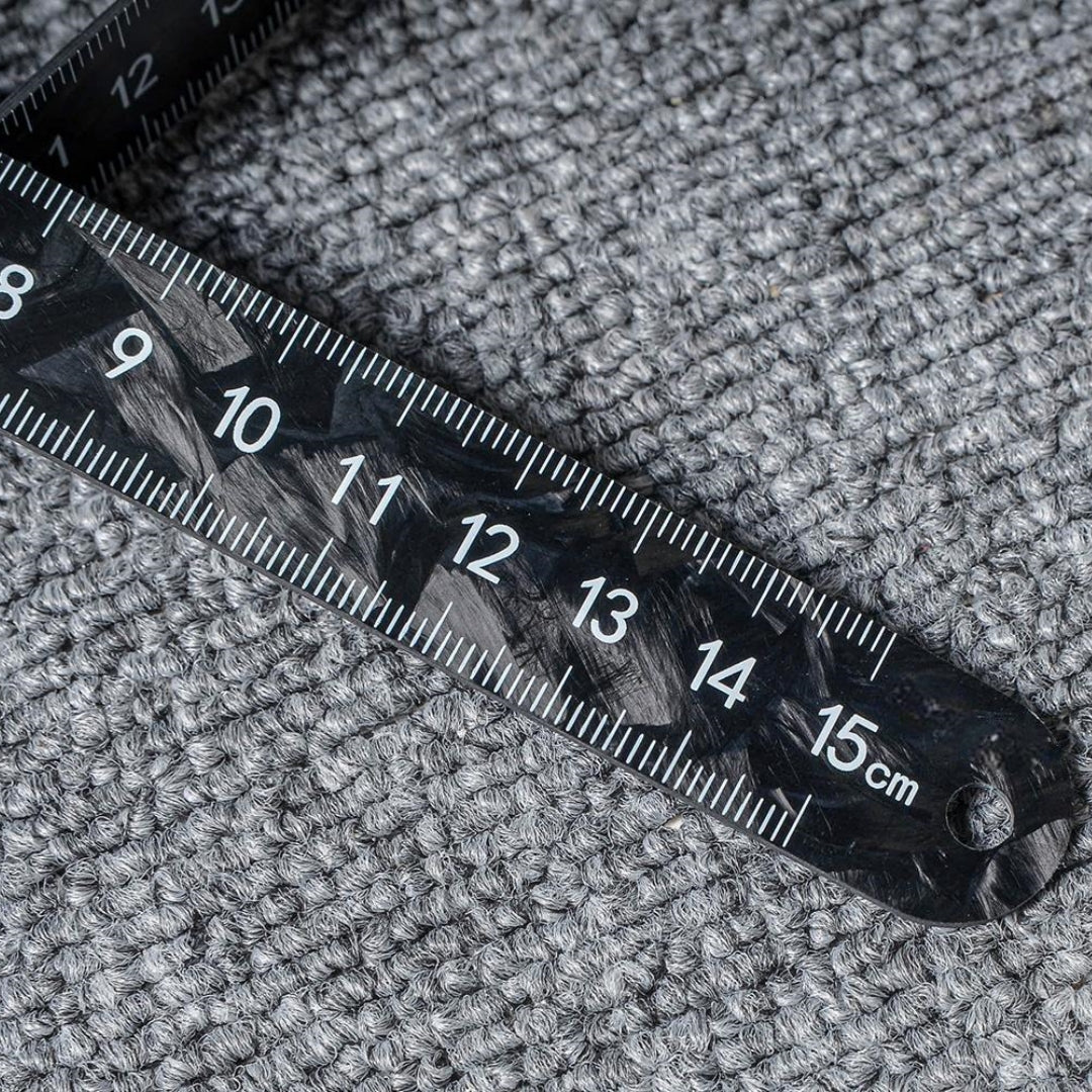 Close-up of a forged carbon fiber ruler with distinct patterns and clear measurement markings up to 15 cm, placed on a textured surface.
