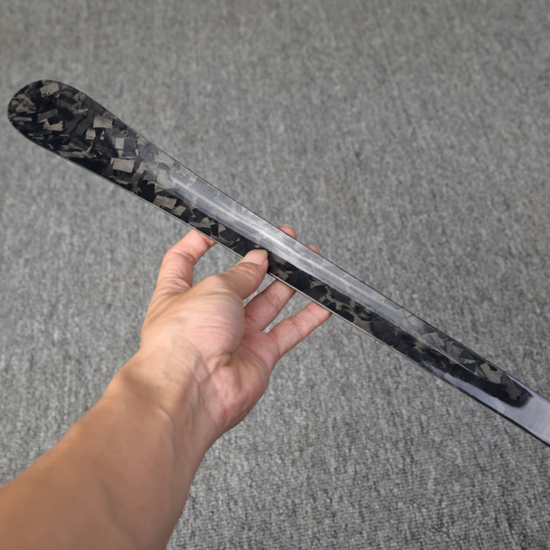 Close-up detail of a long forged carbon fiber shoe horn with a distinct chopped texture, held in a hand against a gray textured surface.
