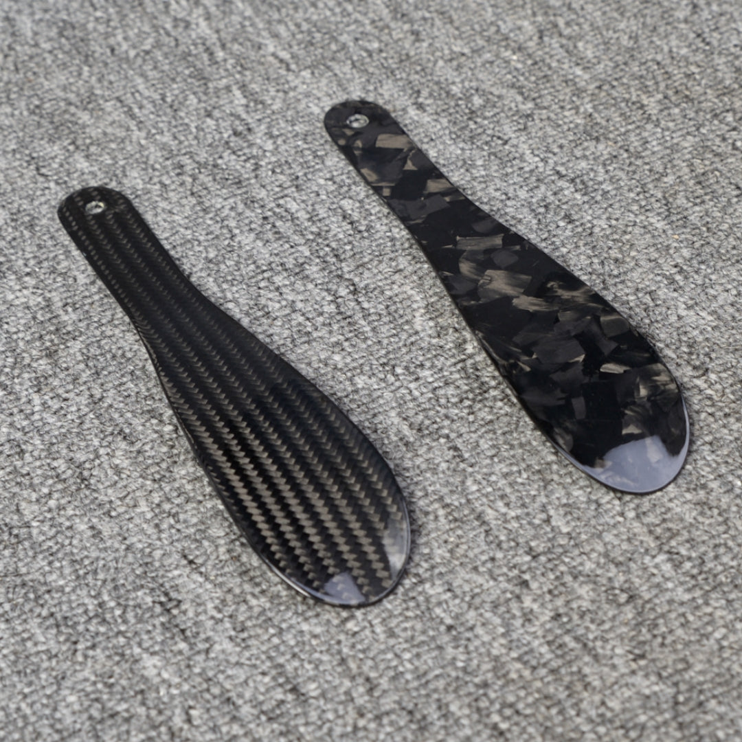 Close-up detail of two short carbon fiber shoe horns—one with a 3K carbon fiber woven pattern and the other with a forged carbon fiber chopped texture—placed on a gray textured surface.
