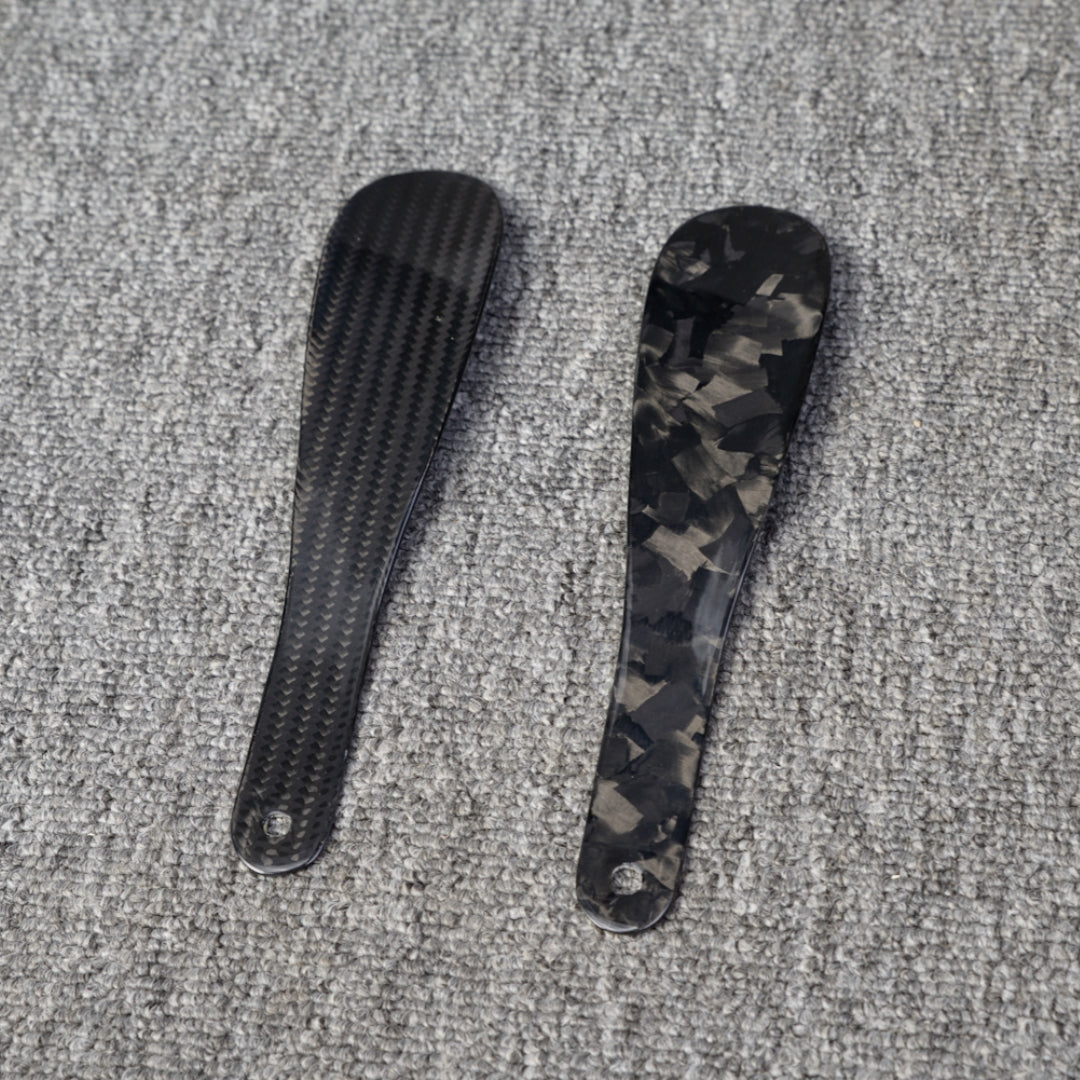 Close-up detail of two short carbon fiber shoe horns—one with a 3K carbon fiber woven pattern and the other with a forged carbon fiber chopped texture—placed on a gray textured surface.