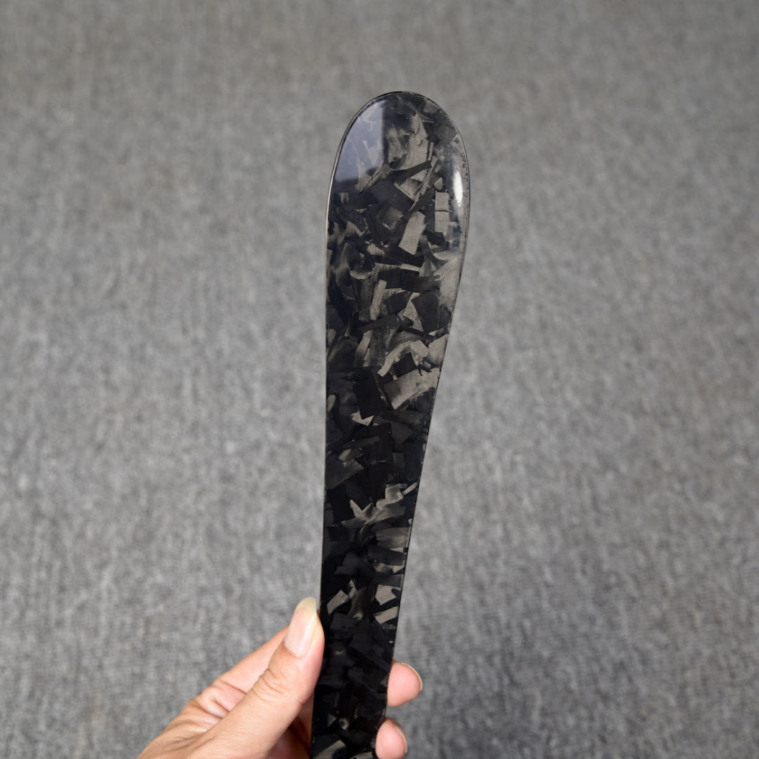 Close-up detail of a long forged carbon fiber shoe horn with a distinct chopped texture, held in a hand against a gray textured background.
