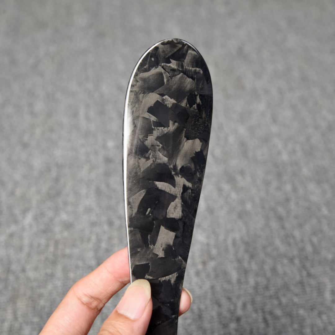 Close-up detail of a short forged carbon fiber shoe horn with a distinct chopped texture, held in a hand against a gray textured background.