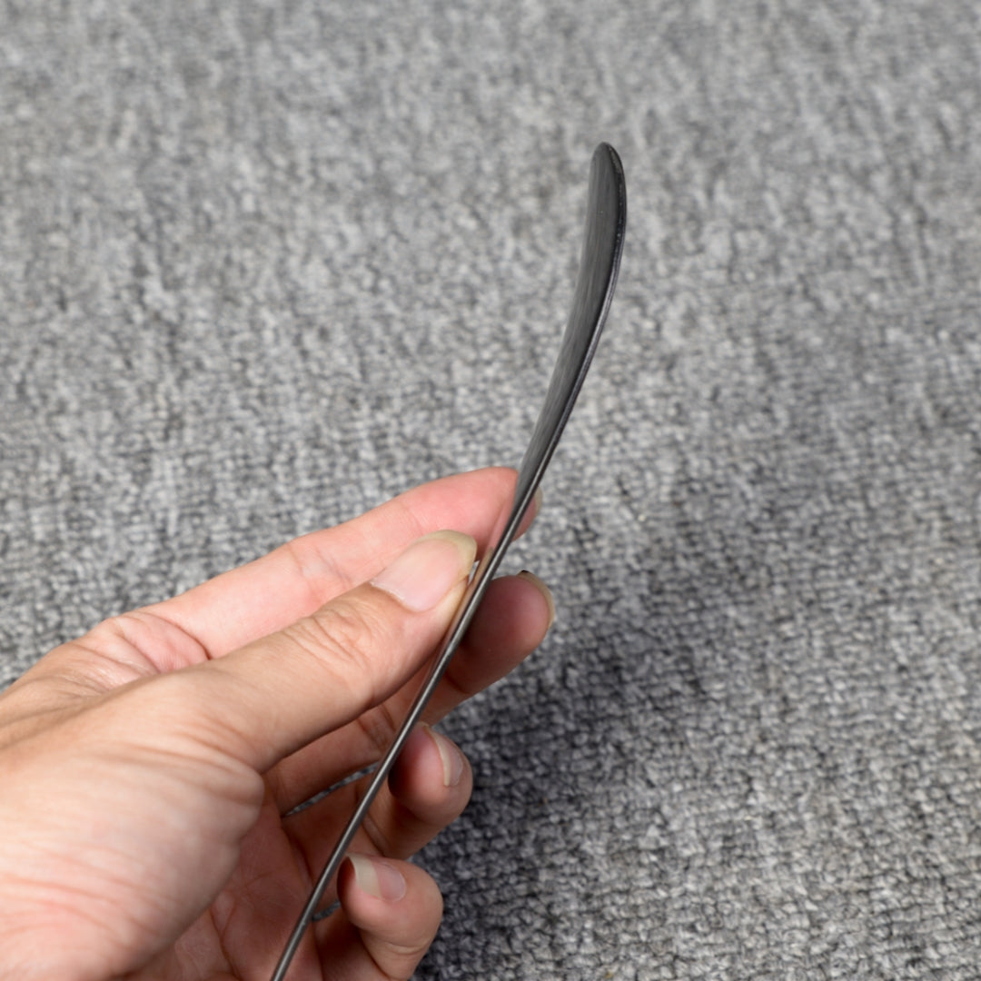 Close-up detail of a short 3K carbon fiber shoe horn with a distinct woven pattern, held in a hand against a gray textured background.