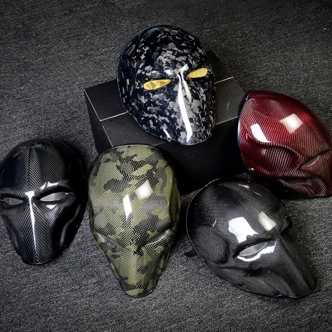 Close-up of multiple carbon fiber tactical masks in various styles — including camouflage, glossy black, and red carbon fiber patterns — arranged on a dark textured surface.