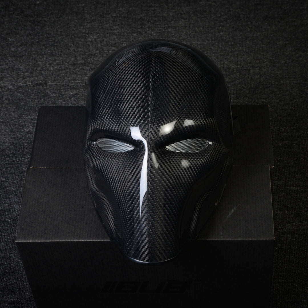 Close-up of a 3K twill carbon fiber tactical mask with mesh eye openings and a glossy finish, displayed on a black box against a dark textured background.