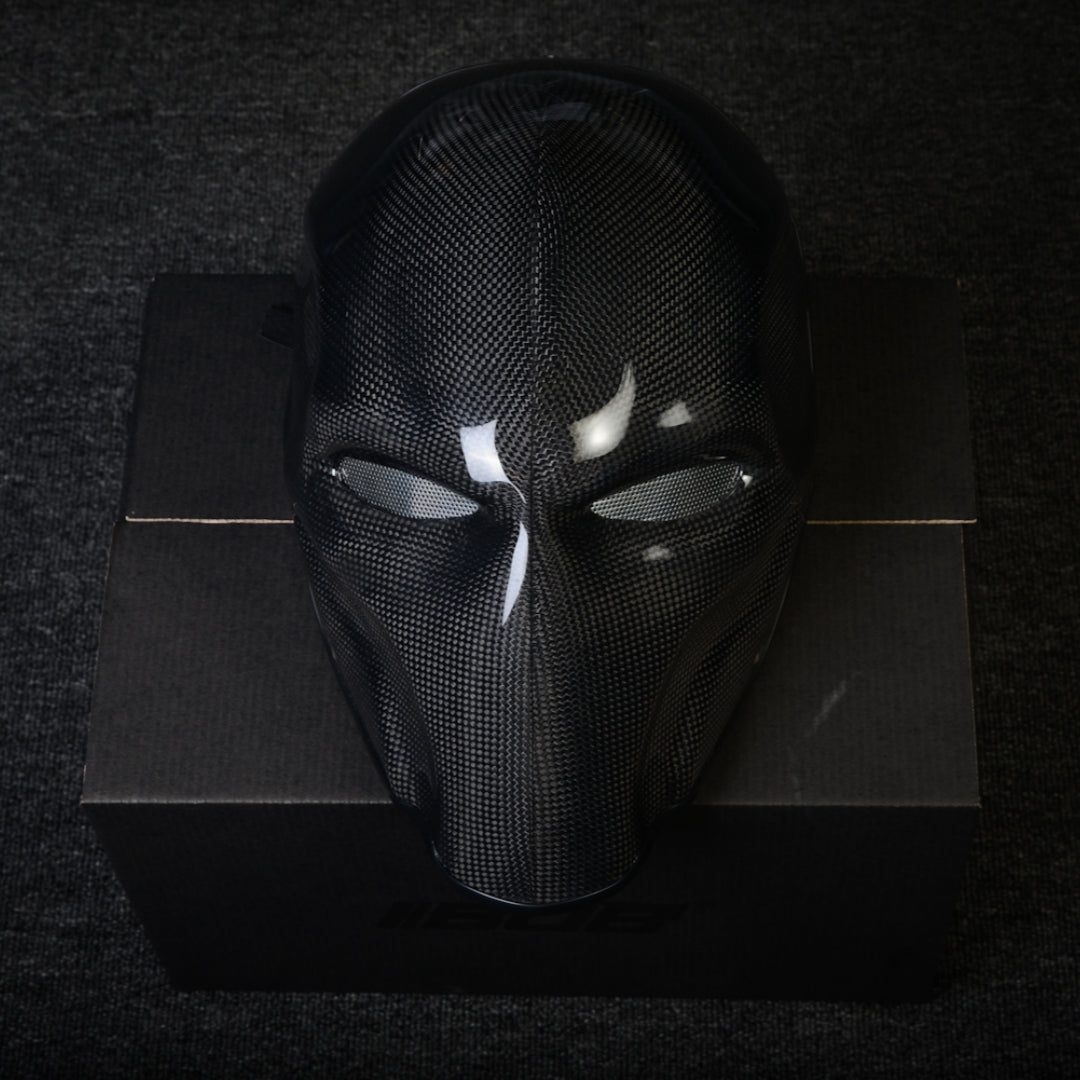 Close-up of a 3K plain weave carbon fiber tactical mask with mesh eye openings and a glossy finish, displayed on a black box against a dark textured background.