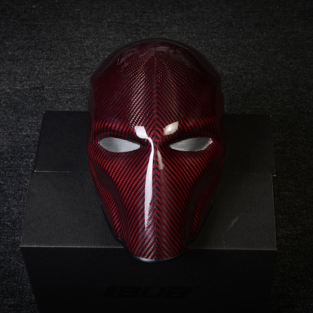 Close-up of a red 3K twill carbon fiber tactical mask with mesh eye openings and a glossy finish, displayed on a black box against a dark textured background.