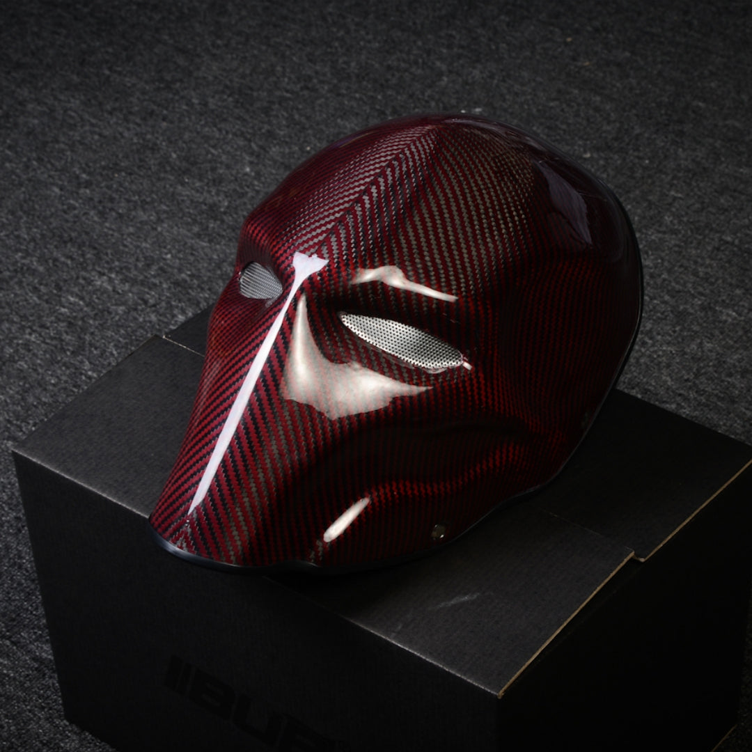 Close-up of a red 3K carbon fiber tactical mask with a glossy finish and mesh eye openings, displayed on a black box against a dark textured background.
