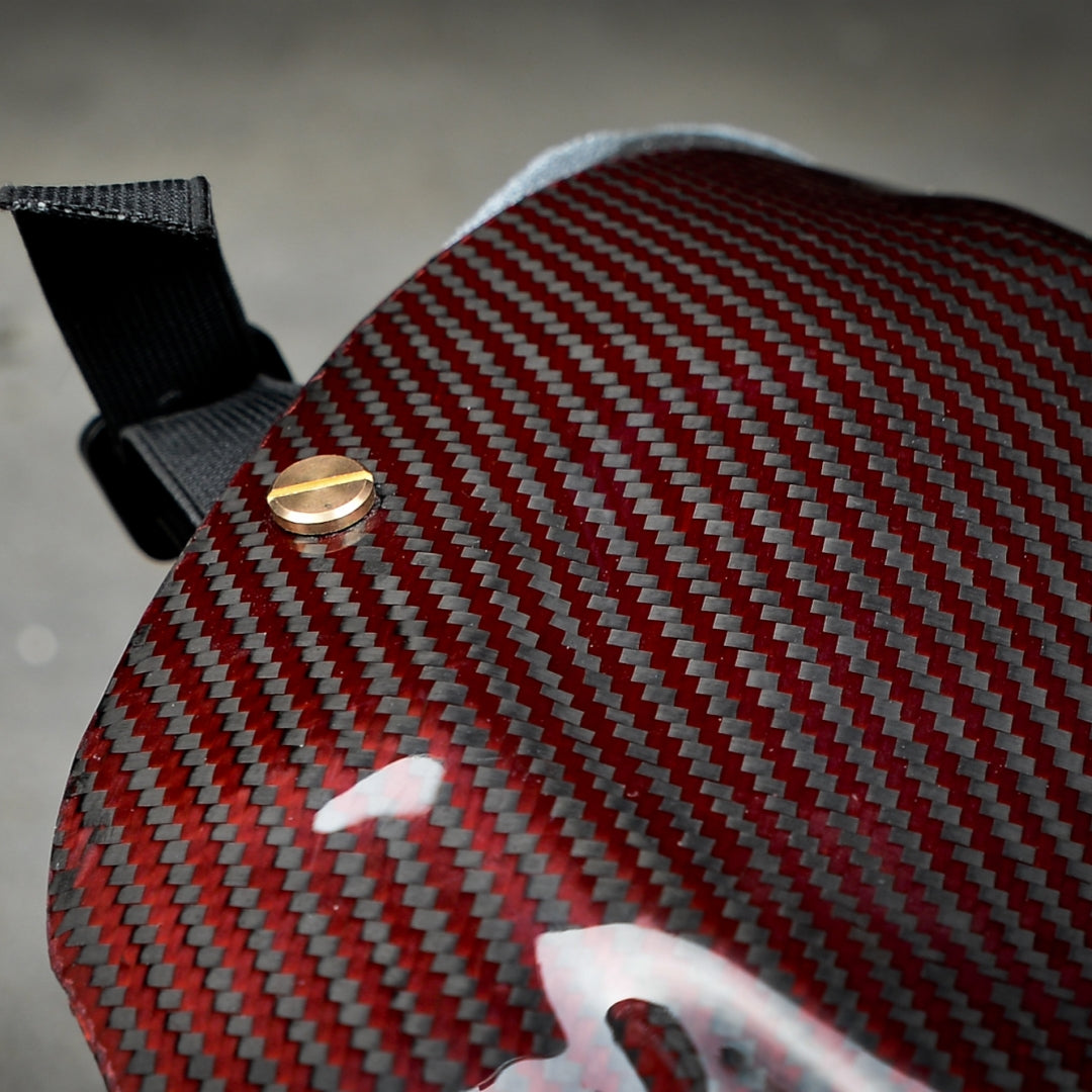 Close-up detail of a red and black 3K carbon fiber tactical mask, showcasing the twill pattern, glossy finish, metal fastener, and black strap.