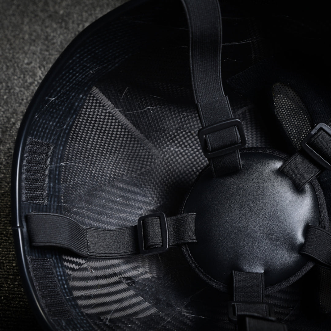 Close-up of the interior of a 3K twill carbon fiber tactical mask, showcasing black adjustable straps with buckle fasteners and the visible carbon fiber pattern through the inner surface, against a textured background.