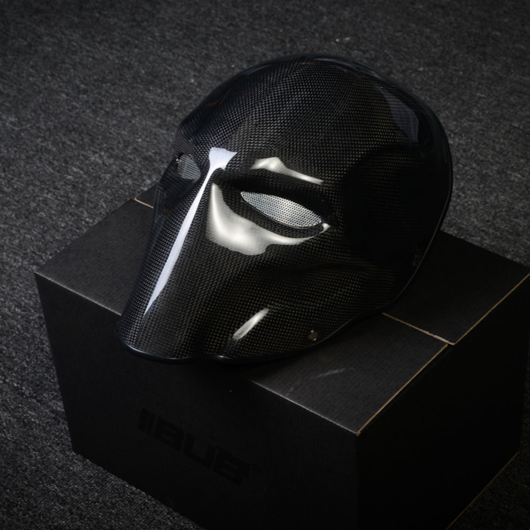 Close-up of a black 3K plain weave carbon fiber tactical mask with a glossy finish and mesh eye openings, displayed on a black box against a dark textured background.