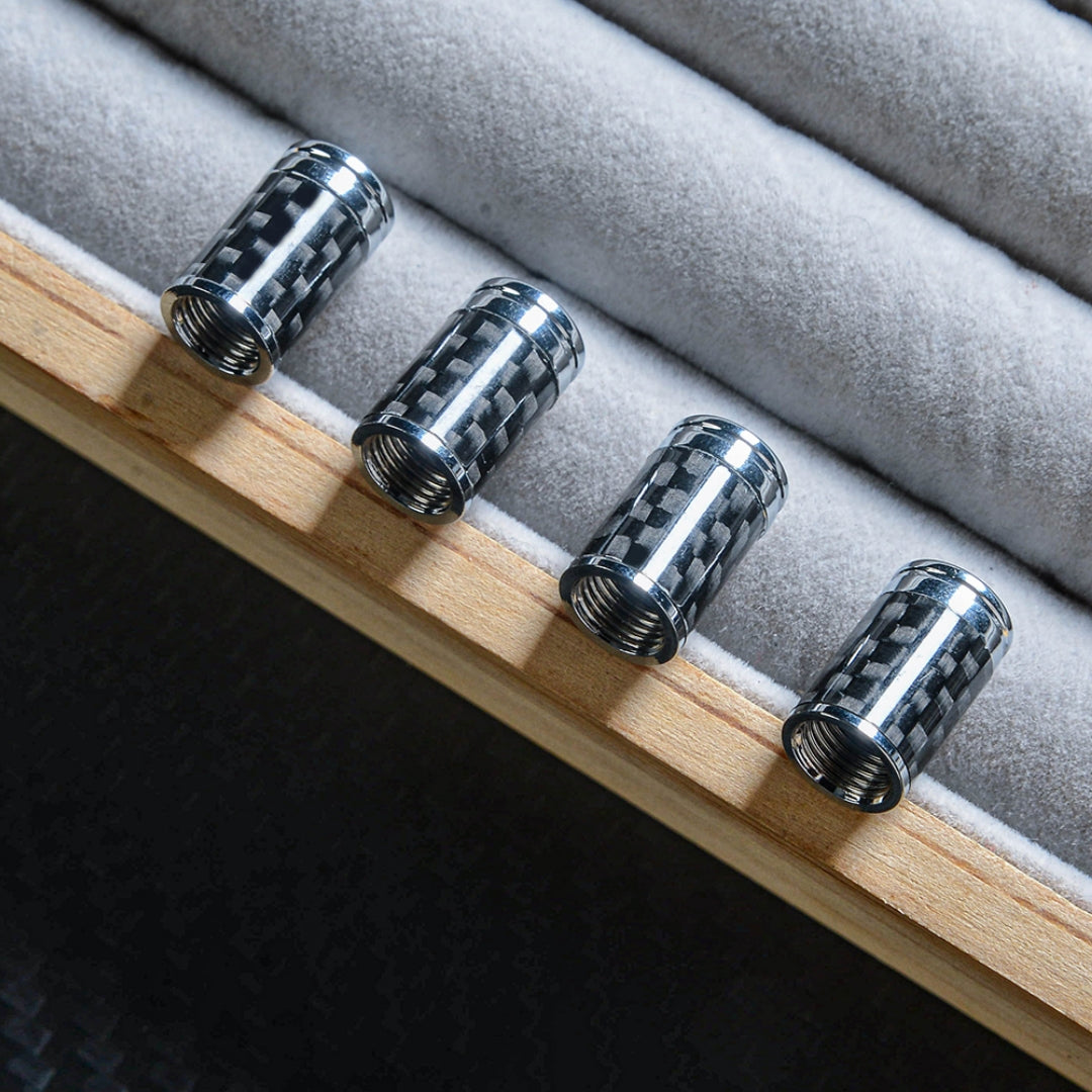Close-up of four carbon fiber car tire valve stem caps with chrome detailing, lined up on a wooden edge with gray velvet padding, showcasing the carbon fiber pattern against a dark carbon fiber-textured background.