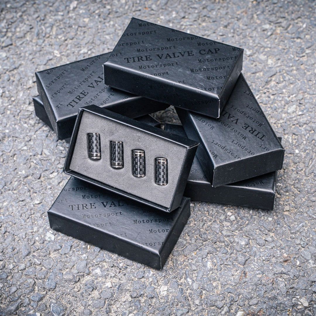 Close-up of four carbon fiber car tire valve caps displayed in an open black box with “TIRE VALVE CAP” and “Motorsport” branding, surrounded by stacked boxes, set against a rough asphalt surface.