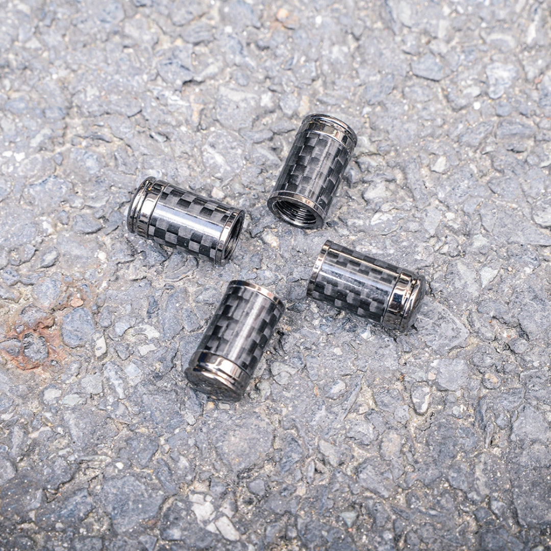 Close-up of four carbon fiber car tire valve stem caps with a checkered pattern and metallic accents, scattered on a rough asphalt surface.