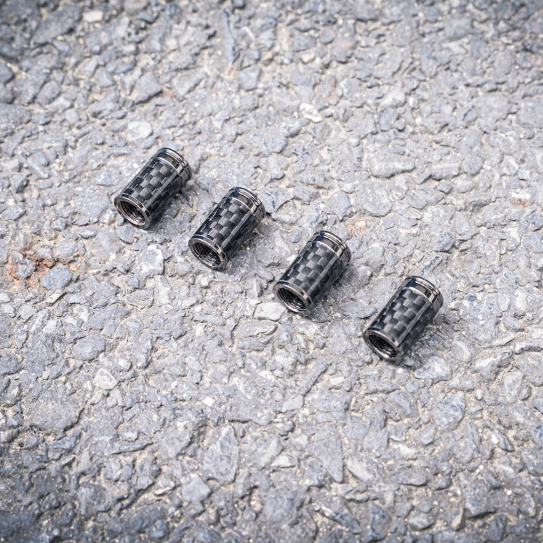 Close-up of four carbon fiber car tire valve stem caps with metallic accents, placed on a rough asphalt surface.