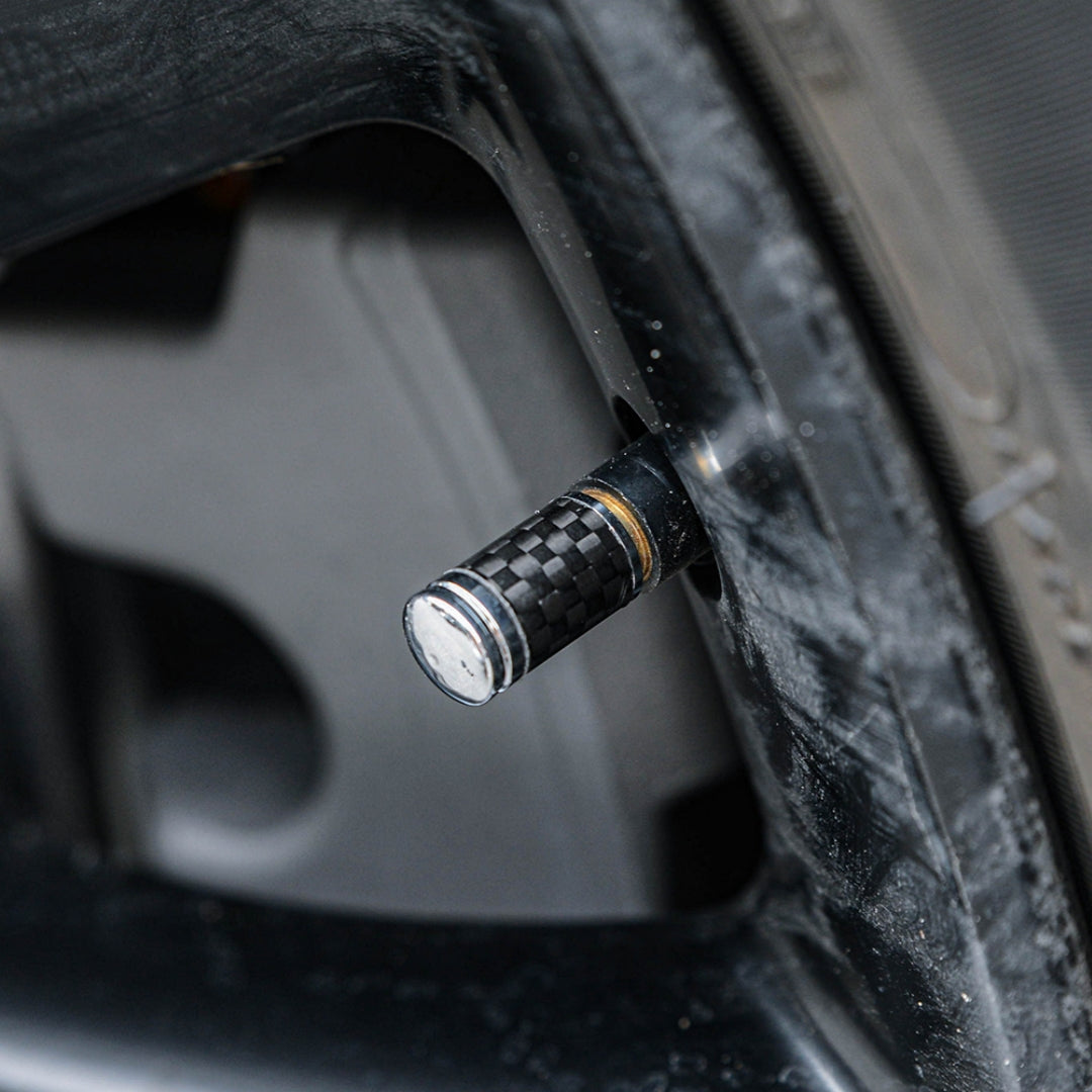 Close-up of a carbon fiber car tire valve stem cap with a checkered pattern and metallic detailing, installed on a vehicle's wheel rim with the tire partially visible in the background.