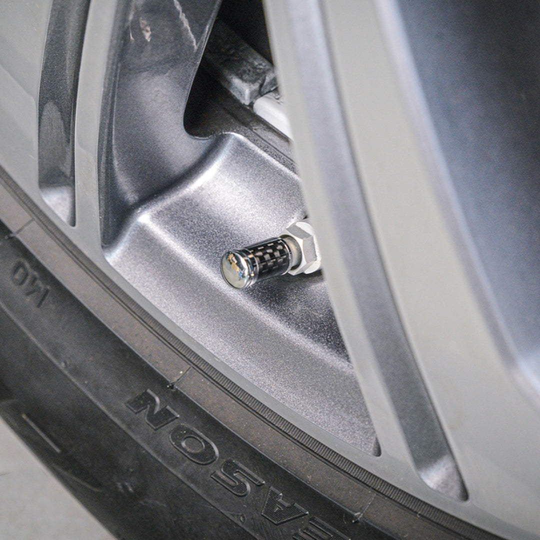 Close-up of a carbon fiber car tire valve stem cap with a checkered pattern, installed on a silver vehicle wheel rim with part of the tire visible.