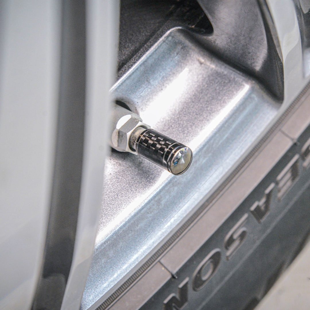 Close-up of a carbon fiber car tire valve stem cap with a checkered pattern, installed on a silver vehicle wheel rim with part of the tire visible.