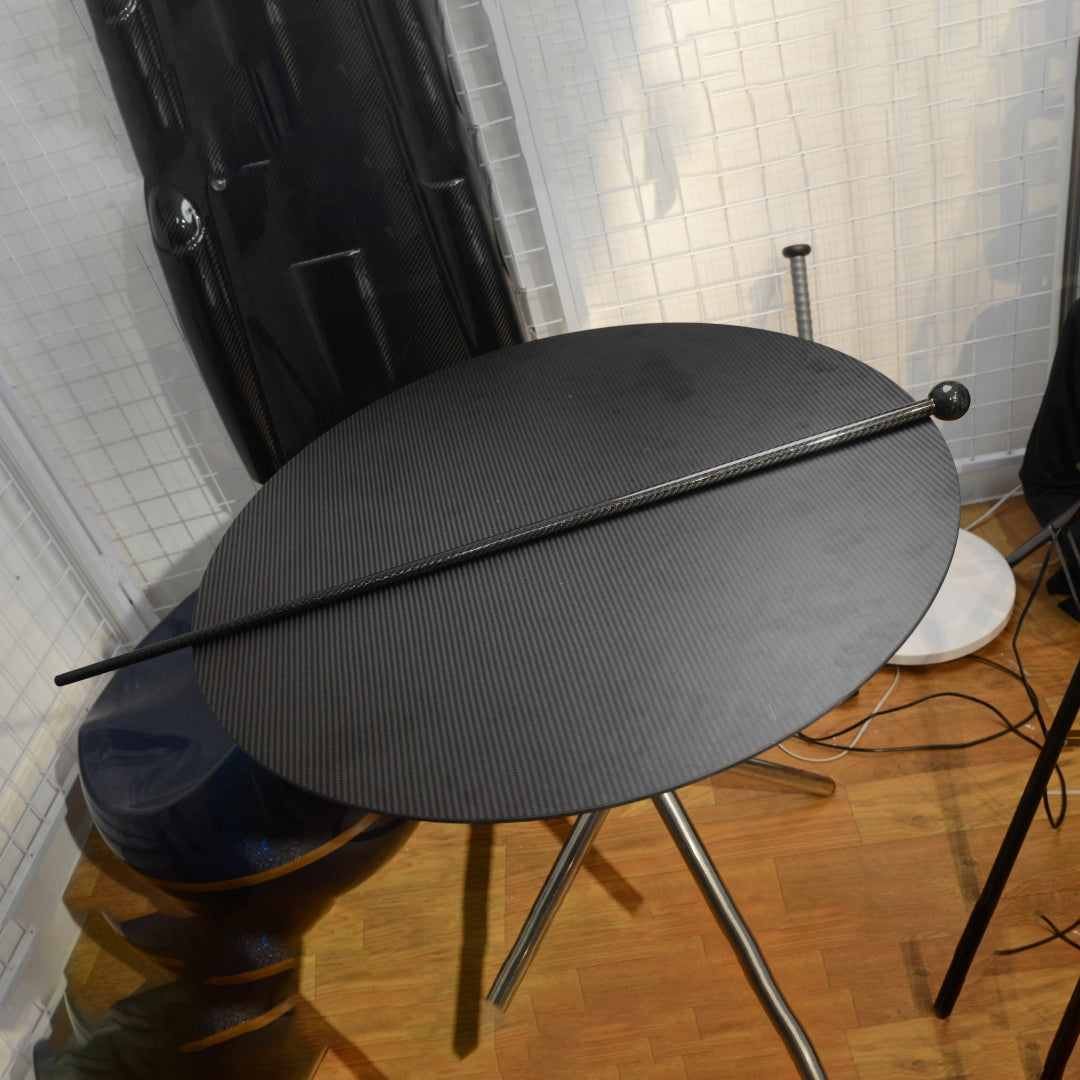 Close-up of a full-length carbon fiber cane with a spherical top, placed on a carbon fiber textured round table, with other carbon fiber items visible in the background.