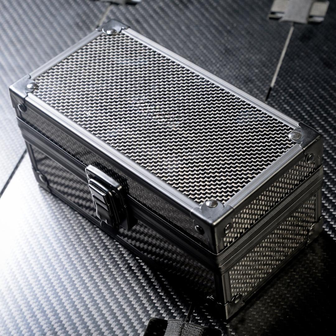 Close-up of a 3K carbon fiber watch box featuring a checkered pattern, metallic corner protectors, and a black latch, positioned on a carbon fiber textured surface.