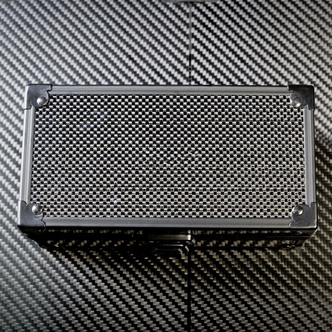 Close-up of a rectangular 3K carbon fiber watch box with a checkered pattern and metallic corner accents, placed on a carbon fiber textured surface.