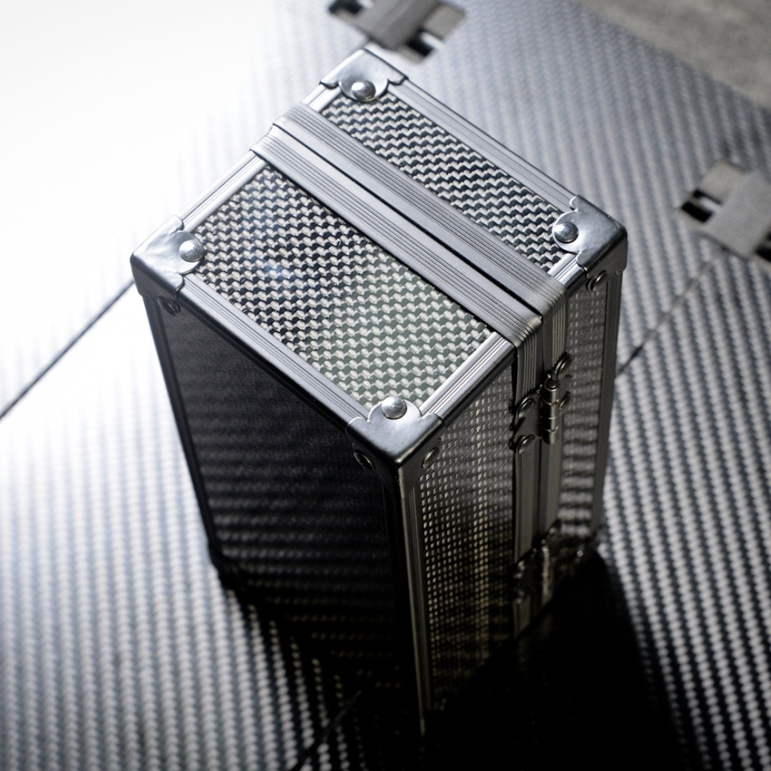 Close-up of a cube-shaped 3K carbon fiber watch box with a checkered pattern and metallic corner reinforcements, positioned on a carbon fiber textured surface.