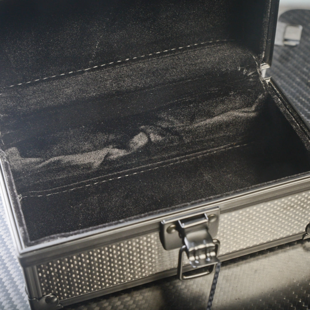 Close-up of an open 3K carbon fiber watch box showcasing a black velvet interior and a metallic latch, with a carbon fiber textured exterior visible.