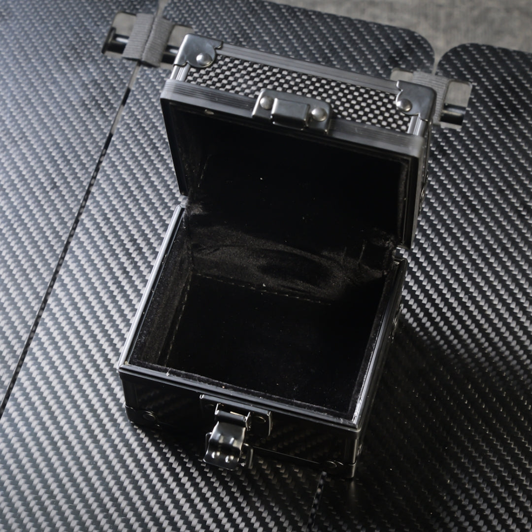Close-up of an open 3K carbon fiber watch box with a black velvet interior, metallic latches, and a carbon fiber textured exterior, placed on a matching carbon fiber surface.