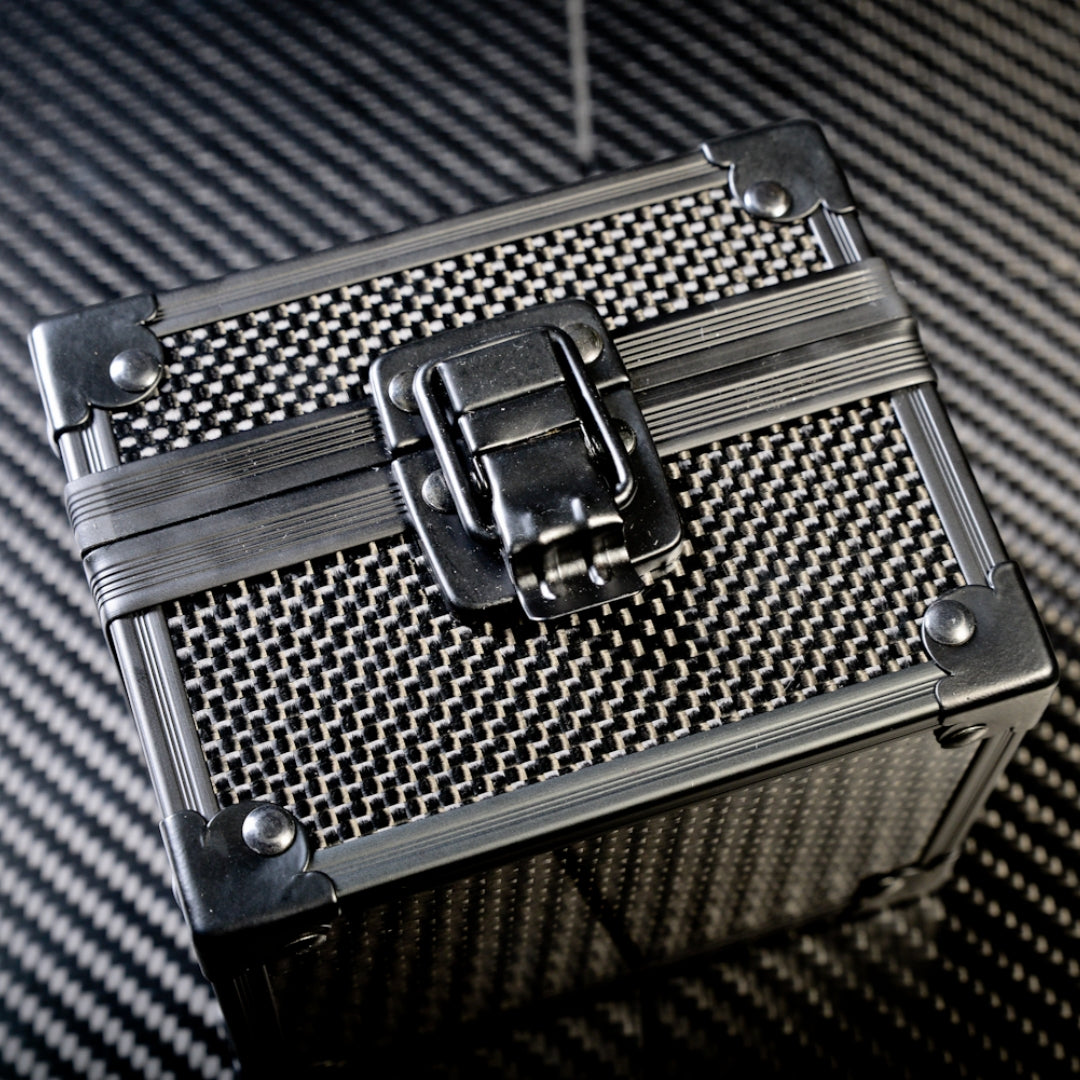 Close-up of a 3K carbon fiber watch box with a checkered pattern, a metallic latch, and reinforced metallic corners, positioned on a carbon fiber textured surface.