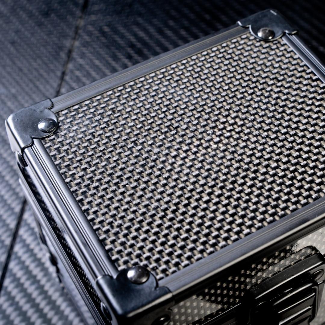 Close-up top view of a 3K carbon fiber watch box featuring a checkered pattern and metallic corner reinforcements, placed on a carbon fiber textured surface.