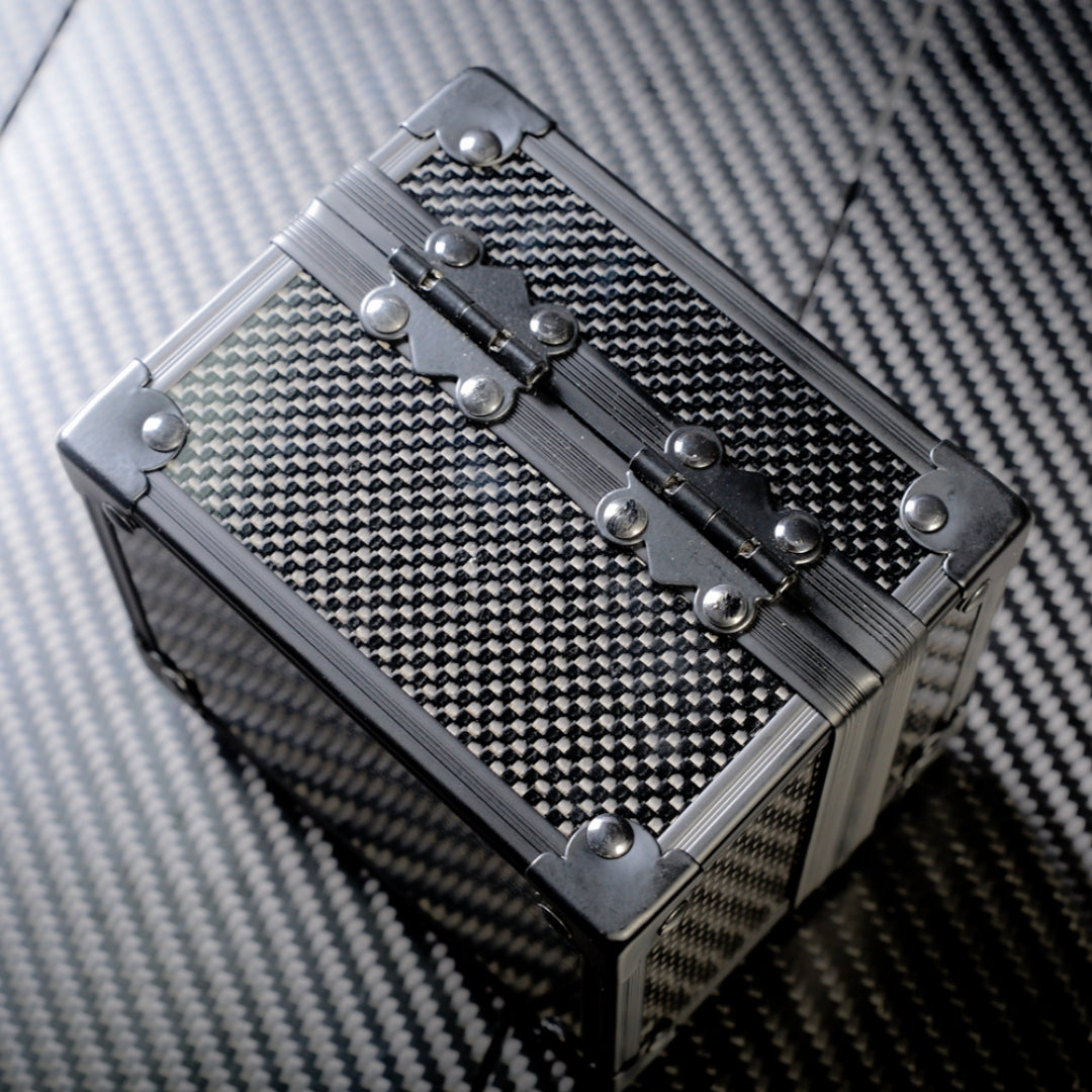 Close-up back view of a 3K carbon fiber watch box with a checkered pattern, metallic hinges adorned with rivets, and reinforced corners, placed on a carbon fiber textured surface.