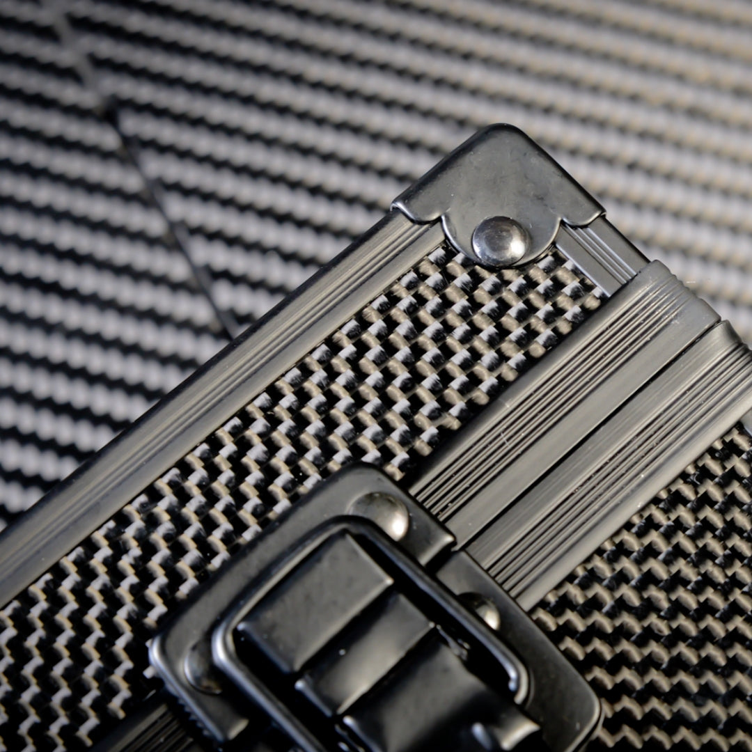 Close-up of the front of a 3K carbon fiber watch box featuring a checkered pattern, a black metallic latch, and metallic rivets with trim, placed on a carbon fiber textured surface.
