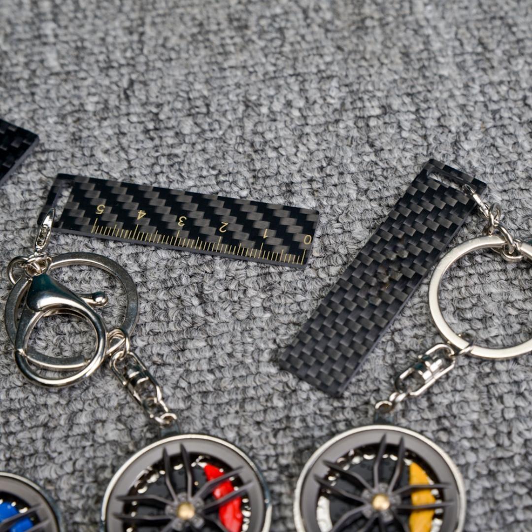 Close-up of two carbon fiber keychains featuring wheel rim designs with red and yellow brake calipers, along with carbon fiber-textured accents (including a ruler-marked piece), displayed on a gray textured fabric surface.