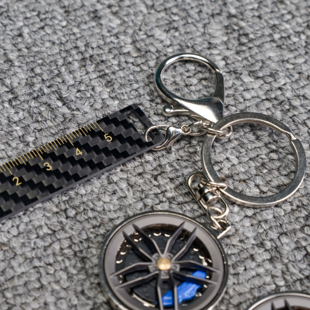 Close-up of a carbon fiber keychain featuring a wheel rim design with blue brake calipers, along with a carbon fiber-textured ruler accent, displayed on a gray textured fabric surface.