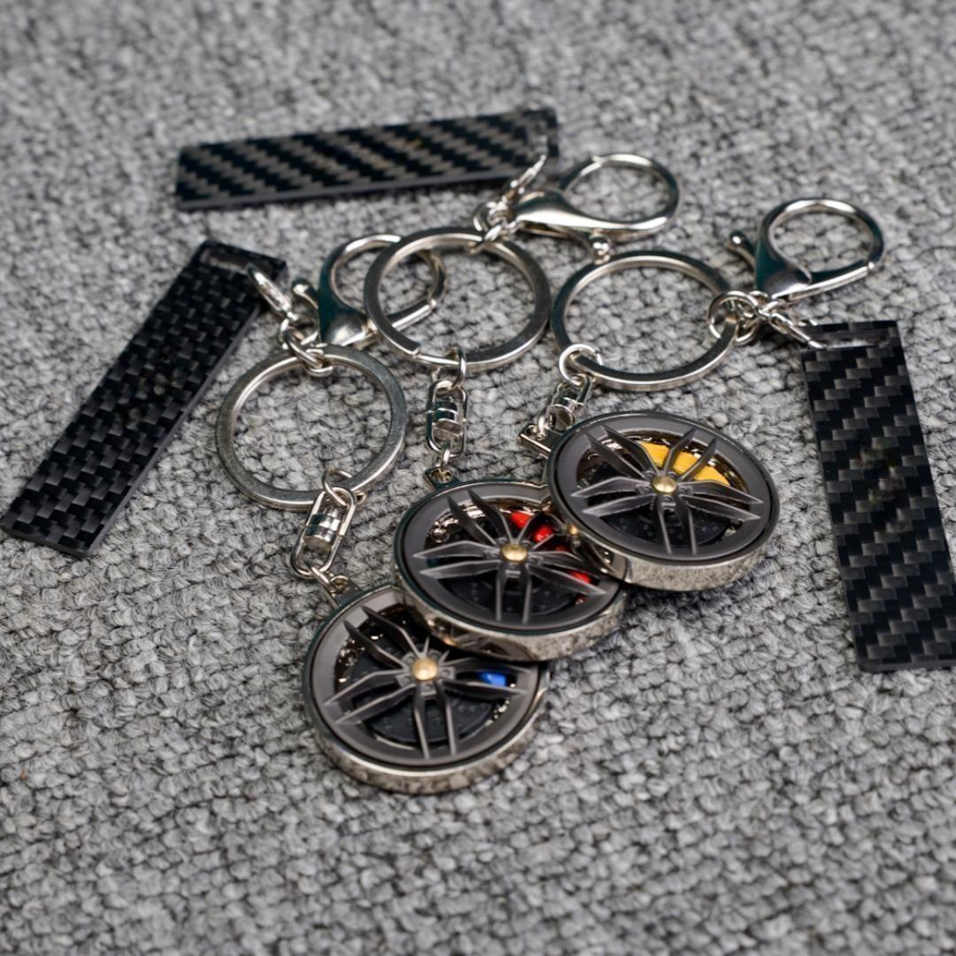 Close-up of three carbon fiber keychains featuring wheel rim designs with colorful brake calipers (blue, red, and yellow), accompanied by carbon fiber-textured accent strips, displayed on a gray textured fabric surface.