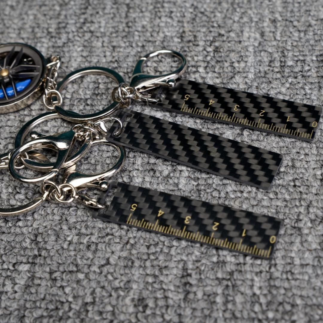 Close-up of three carbon fiber keychains featuring ruler-shaped accents with measurement markings, carbon fiber decorative strips, and a wheel rim charm with blue brake calipers, displayed on a gray textured fabric surface.