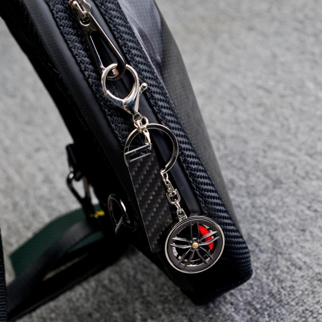 Close-up of a carbon fiber keychain with a wheel rim charm featuring red brake calipers and a carbon fiber-textured accent strip, attached to a black textured bag, displayed on a gray fabric surface.