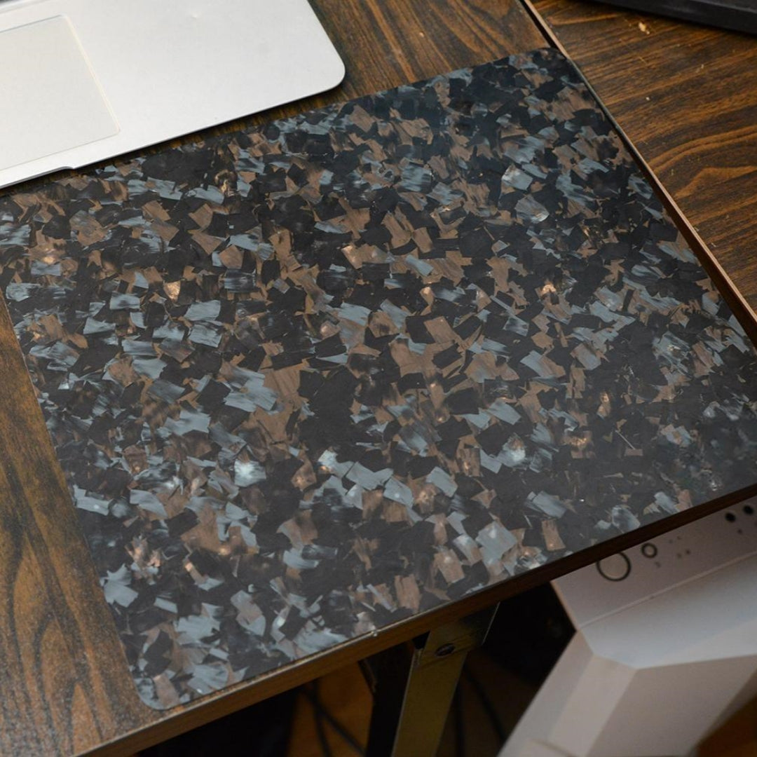 "A forged carbon fiber mouse pad with a textured, fragmented black and brown pattern, placed on a wooden desk with a partial view of a laptop in the background."