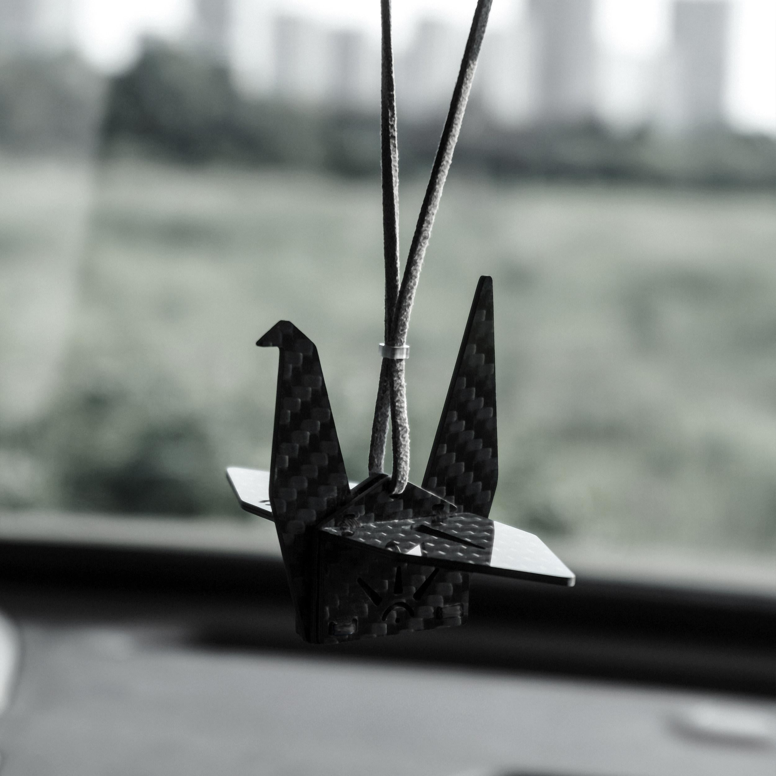 "Black carbon fiber-textured origami crane car charm, hanging from a dark cord inside a vehicle (with a softly blurred outdoor landscape in the background); minimalist modern decorative accessory"