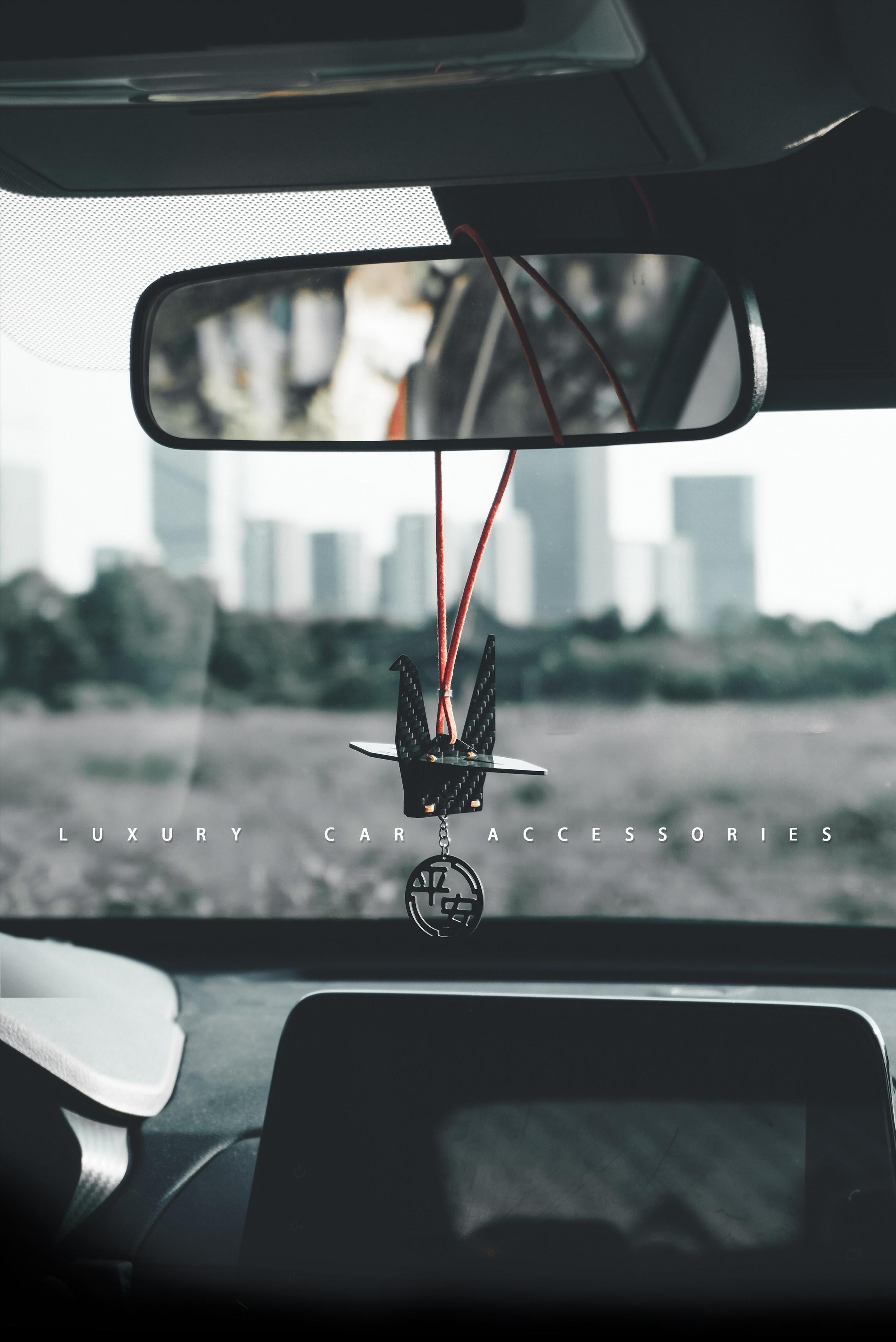 "Luxury car accessory: Black carbon fiber-textured origami crane charm (with small gold accents) hanging from a red cord on a car’s rearview mirror; includes a dangling circular pendant engraved with Chinese 'peace' characters, set against a blurred urban landscape (text overlay reads 'LUXURY CAR ACCESSORIES')"