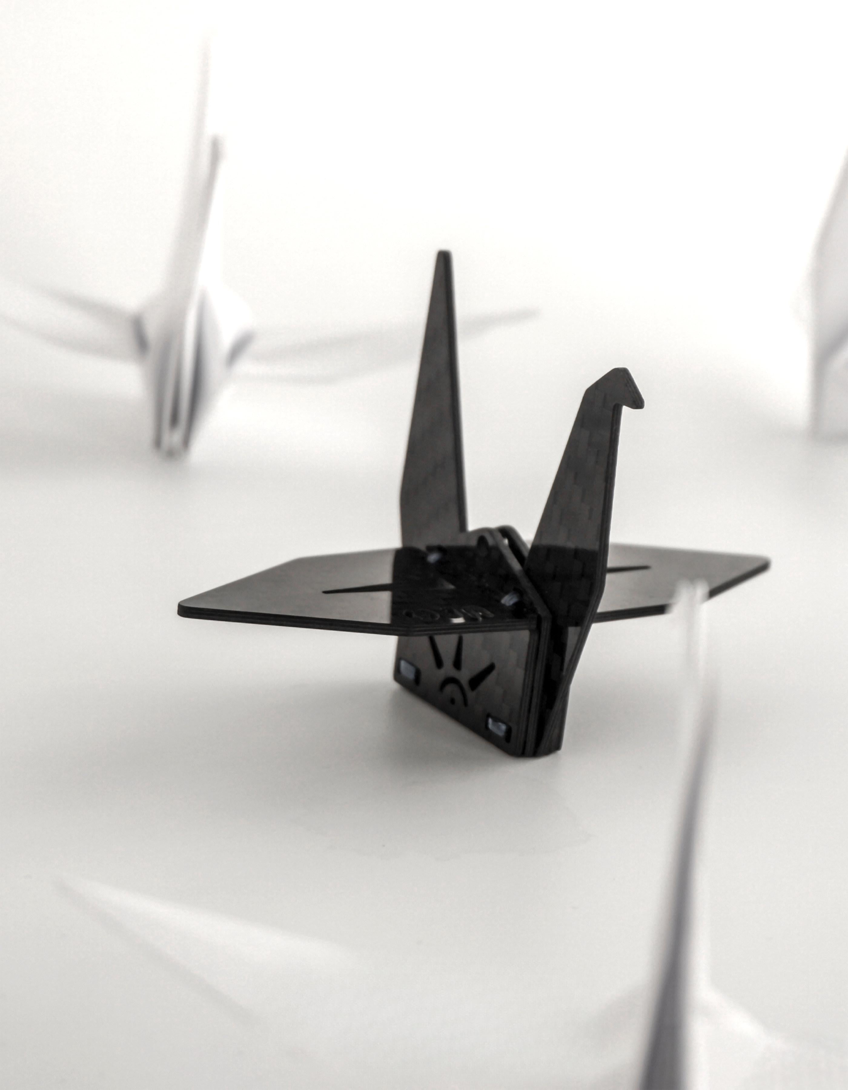"Black carbon fiber-textured origami crane decor (featuring a sun-like design and small square accents on its base), displayed against a plain white surface; softly blurred white origami cranes appear in the background"