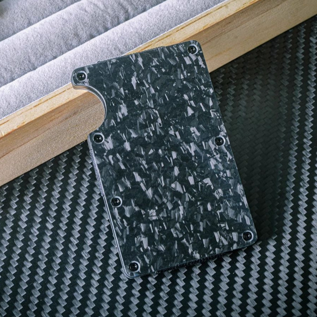 Close-up of a forged carbon fiber card holder with a distinct marbled texture, secured by screw fasteners and featuring a notched design for easy card access, placed on a carbon fiber-textured surface with wooden and soft-lined accents in the background.