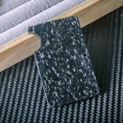 Close-up of a forged carbon fiber card holder with a distinct marbled texture, secured by screw fasteners and featuring a notched design for easy card access, placed on a carbon fiber-textured surface with wooden and soft-lined accents in the background.