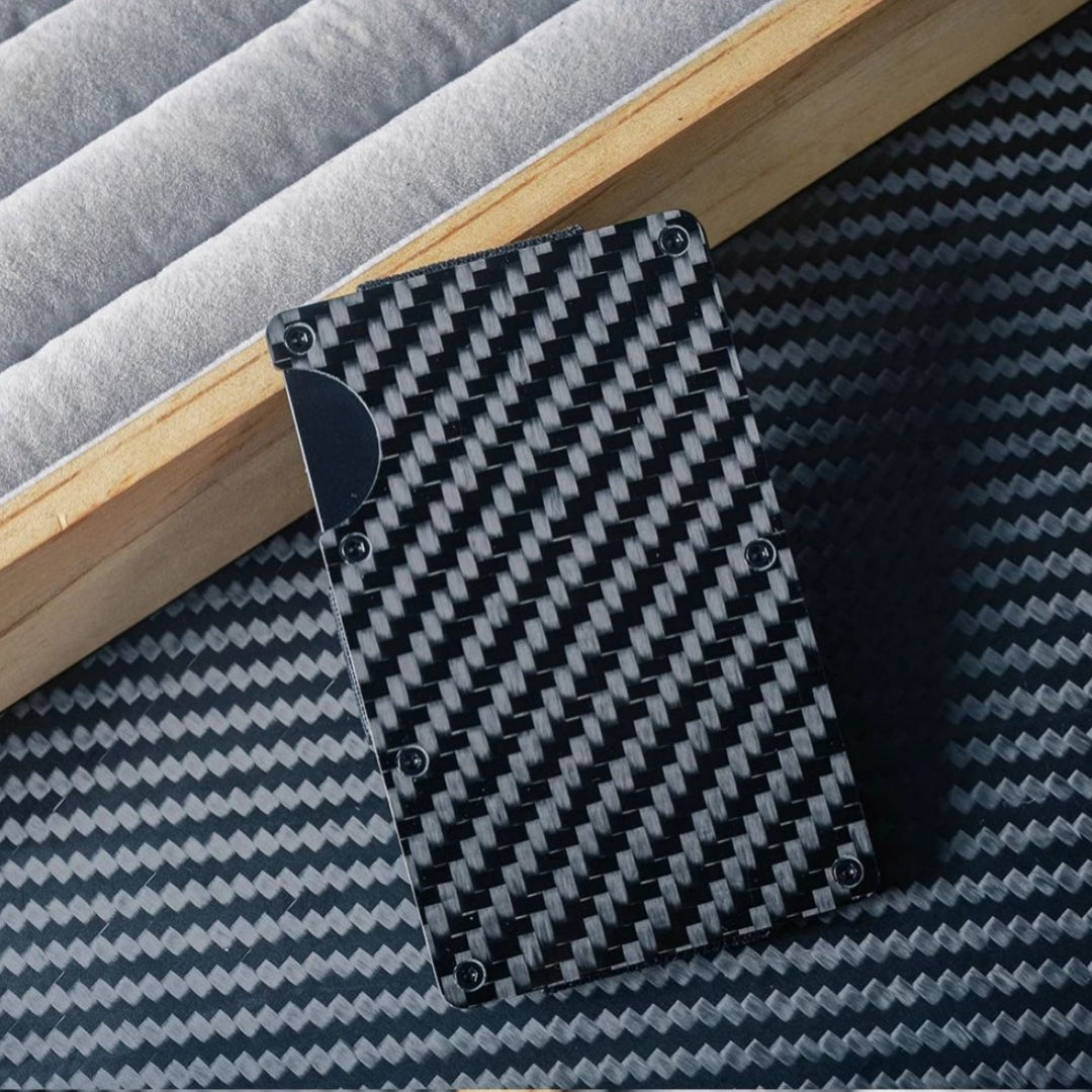 Close-up of a 3K carbon fiber card holder with a distinct woven pattern, secured by screw fasteners and featuring a notched design for easy card access, placed on a carbon fiber-textured surface with wooden and soft-lined accents in the background.