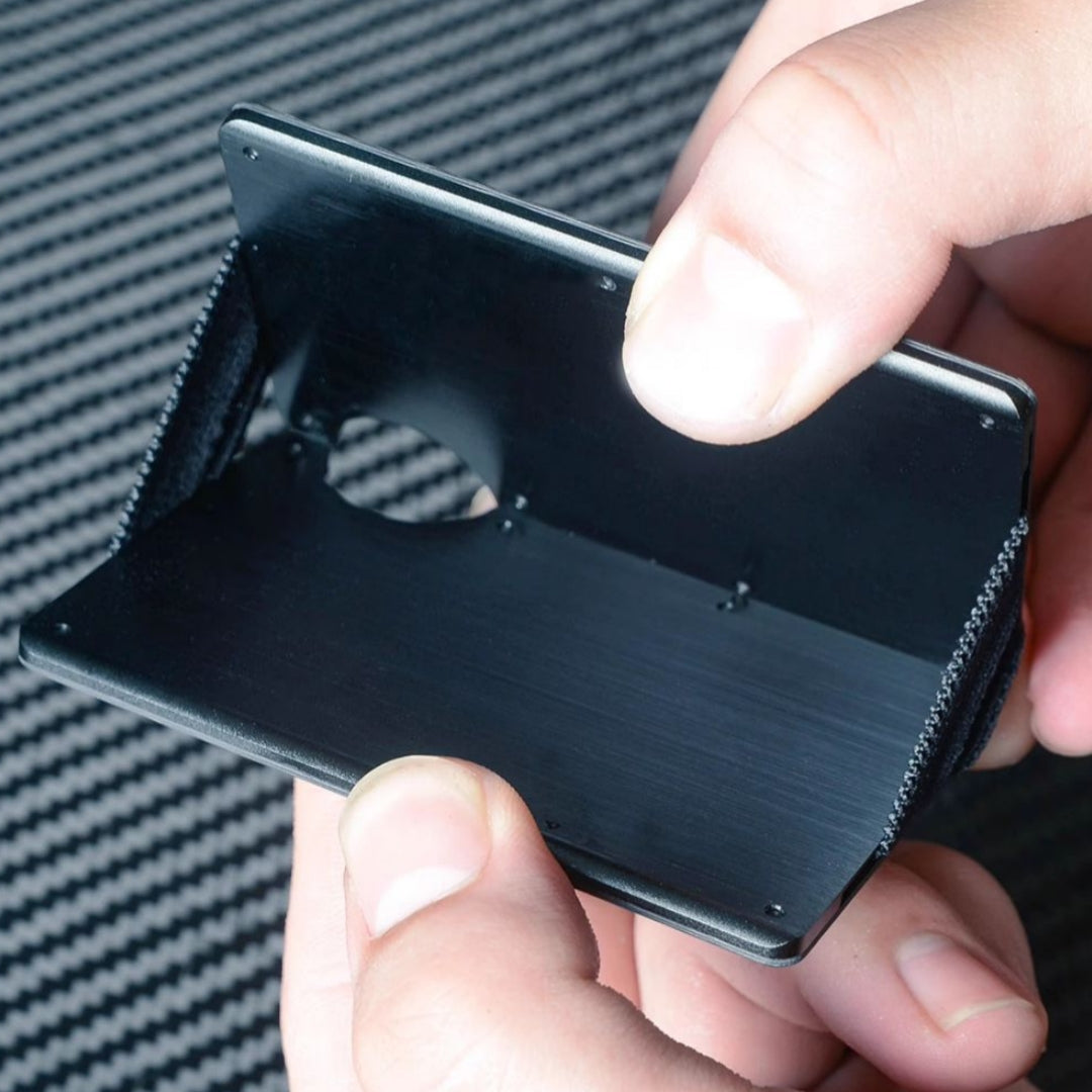 Close-up of hands holding open a 3K carbon fiber card holder, showcasing its interior with a smooth black finish and notched design, against a carbon fiber-textured background.