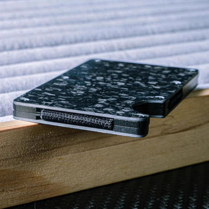 Close-up side view of a forged carbon fiber card holder with a distinct marbled texture, showcasing its closure featuring a hook-and-loop fastener, placed on a wooden surface against a soft, ribbed background.