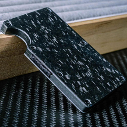Close-up of a forged carbon fiber card holder with a distinct marbled texture, featuring screw fasteners and webbed strap detail on the side, placed on a wooden surface against a carbon fiber-textured background.