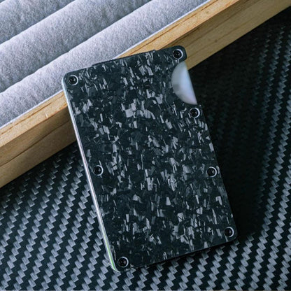 Close-up of a forged carbon fiber card holder with a distinct marbled texture, secured by screw fasteners and featuring a notched design for easy card access, placed on a carbon fiber-textured surface with wooden and soft-lined accents in the background.